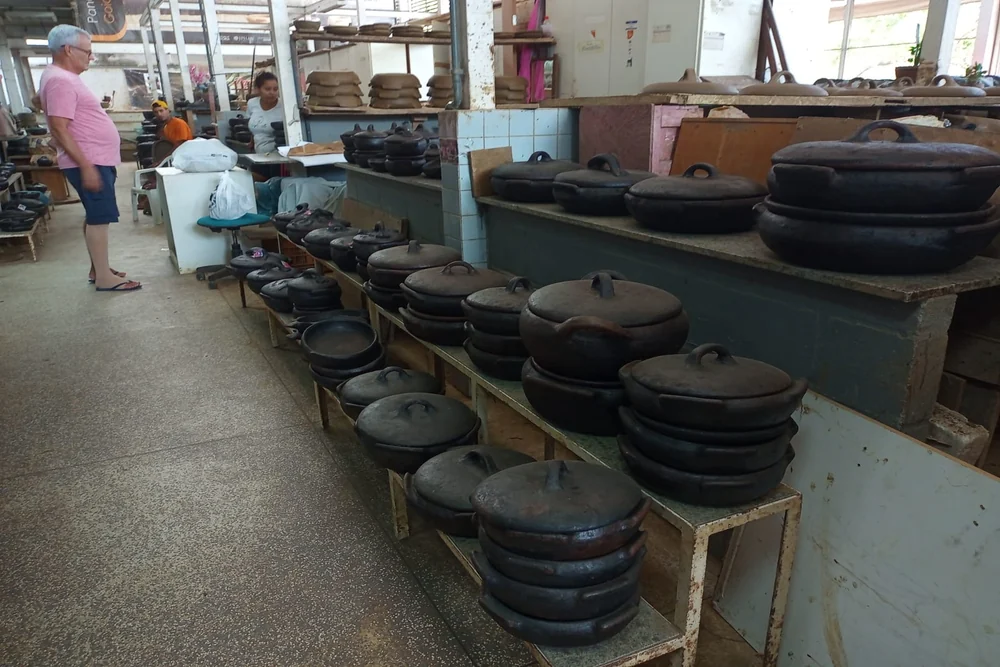 Panelas de barro prontas enfileiradas no galpão da Associação das Paneleiras de Goiabeiras, em Vitória, com artesãs trabalhando ao fundo