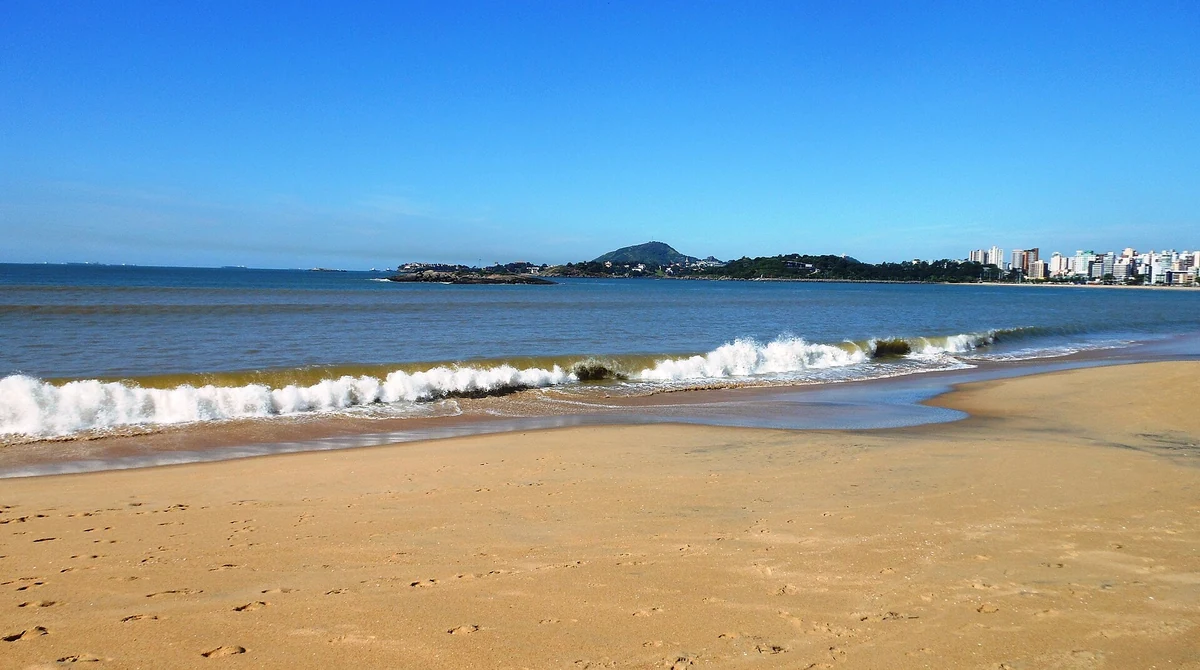 Praia de Camburi, em Vitória - ES, com onda quebrando na areia e a orla da cidade ao fundo