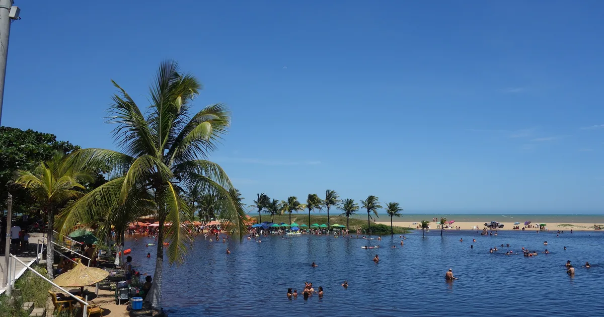 Lagoa de água doce cercada de coqueiros, com o mar ao fundo, em Marataízes - ES
