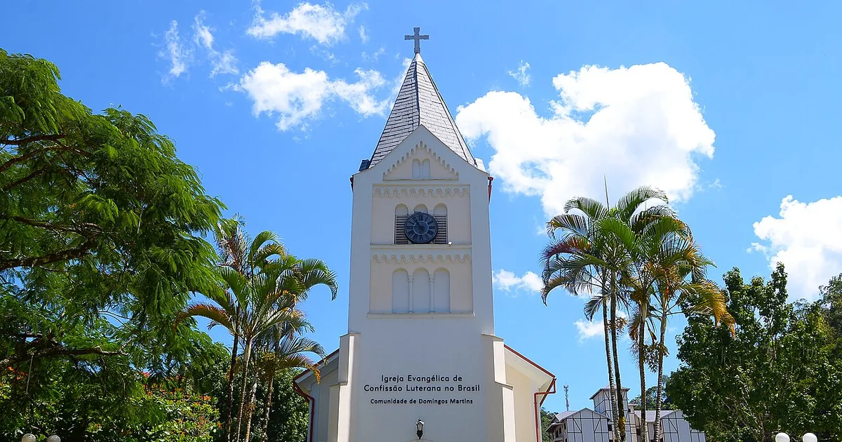 Igreja luterana de torre pontiaguda e relógio, cercada de palmeiras, no centro de Campinho