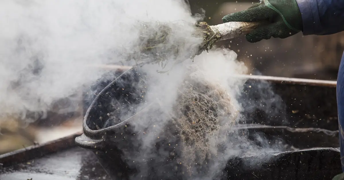 Paneleira aplicando o açoite de tanino em panela de barro quente, com fumaça subindo da peça recém-queimada