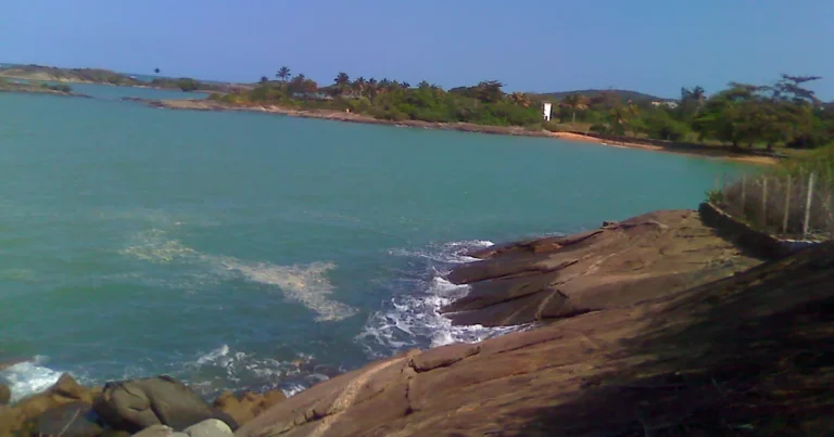 Enseada de água esverdeada entre costões de pedra nas Três Praias, em Guarapari - ES, com coqueiros ao fundo
