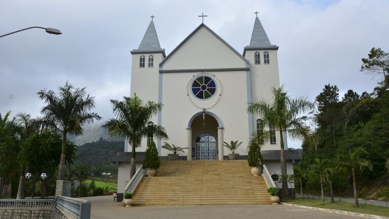 Igreja de fachada branca com duas torres em Domingos Martins, com escadaria, palmeiras e a serra ao fundo