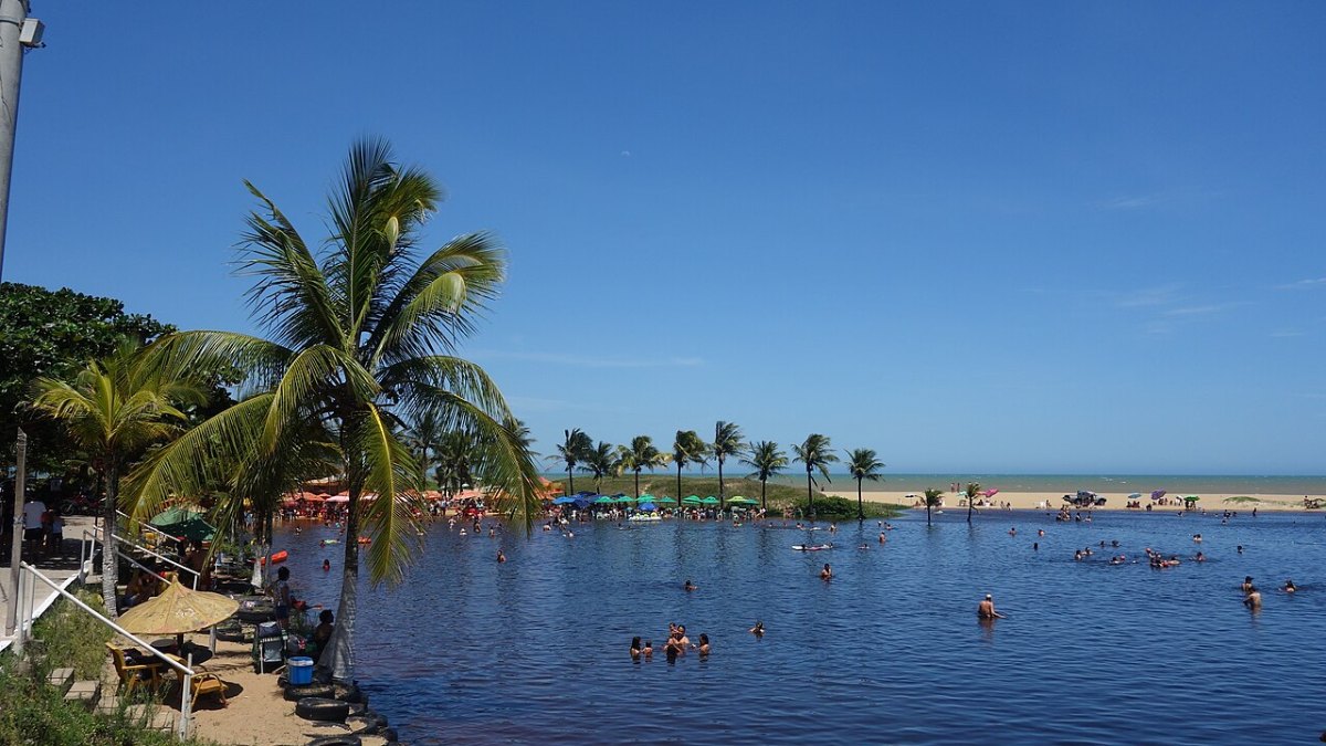 Lagoa de água parada cercada de coqueiros, com faixa de areia e o mar ao fundo, em Marataízes - ES