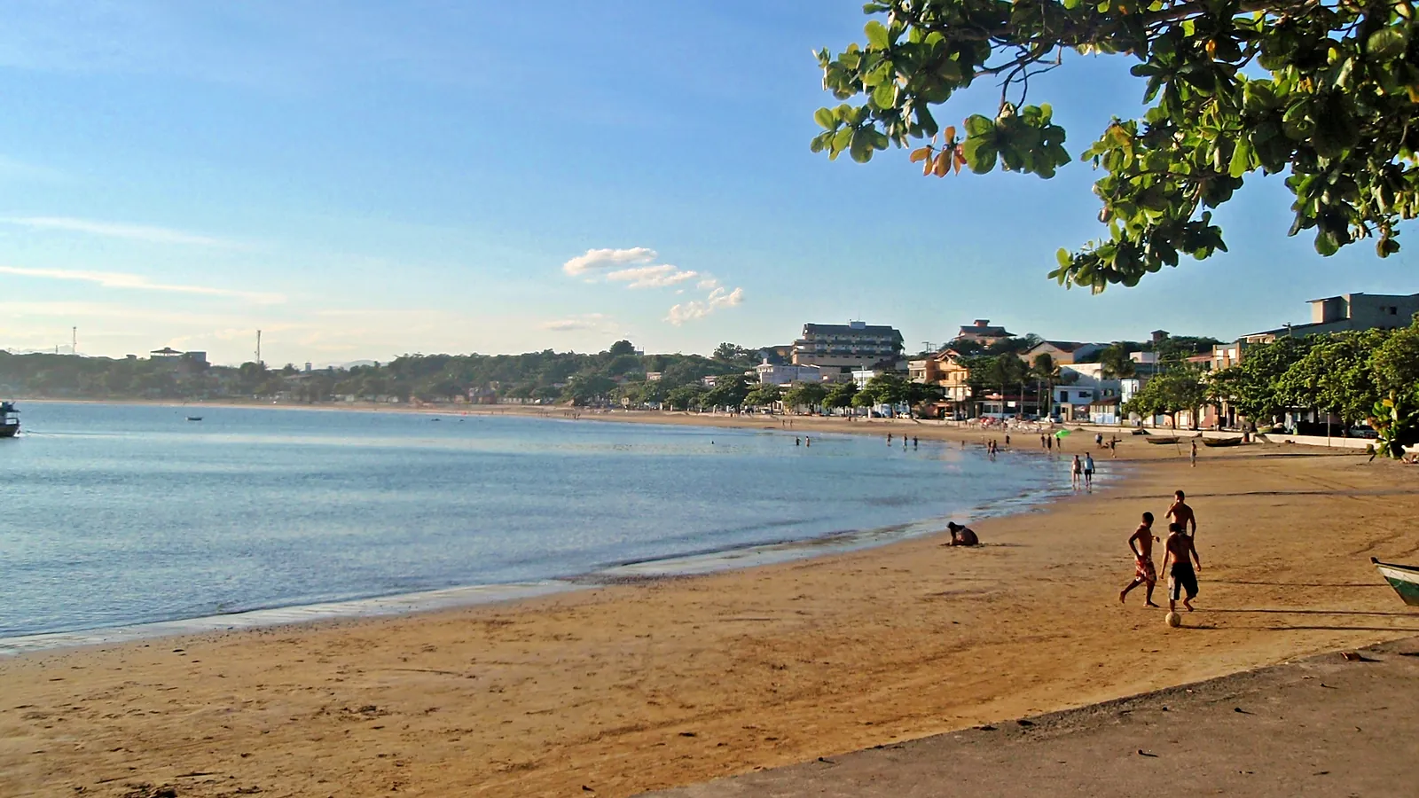 Praia de Ubu em Anchieta (ES) no fim da tarde, com a faixa de areia em curva, o mar calmo, meninos jogando bola e casas e prédios baixos ao fundo