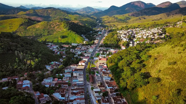 Vista aérea do centro de Muqui (ES), com o casario e a rua principal no fundo do vale entre morros verdes e cafezais, e serras ao fundo sob céu claro de fim de tarde