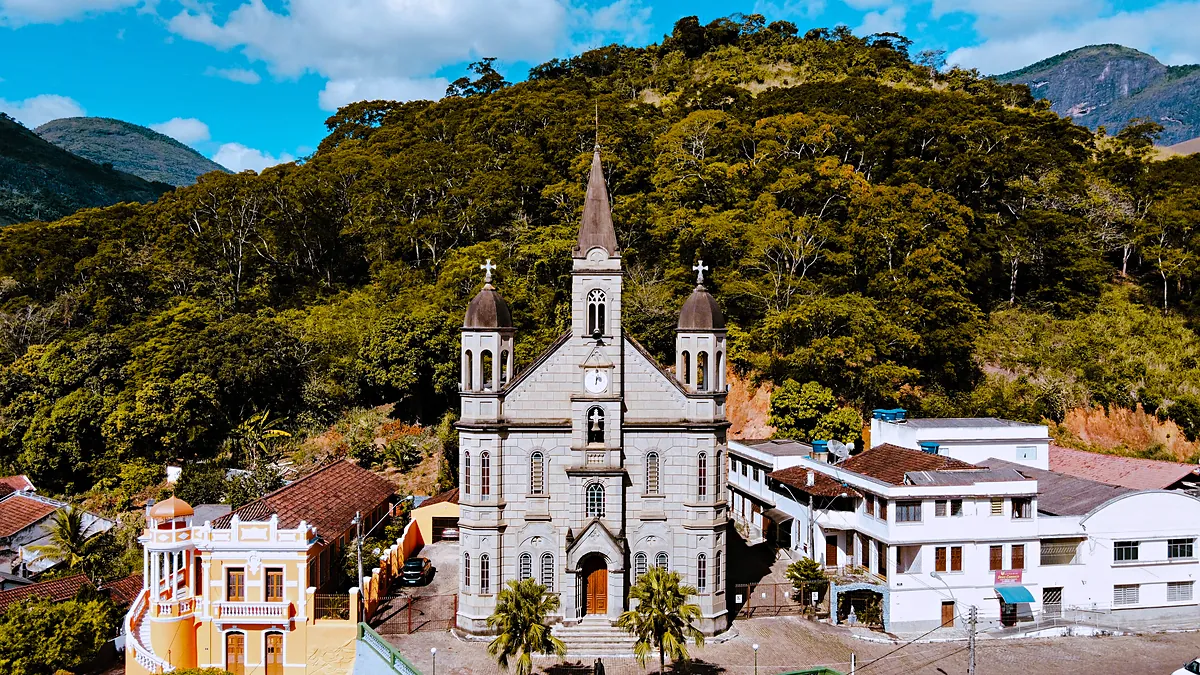 Igreja Matriz de São João Batista vista do alto no centro de Muqui (ES), com fachada de pedra cinza, torre central pontiaguda e duas torres laterais com cúpula, um sobrado amarelo ao lado e morros cobertos de mata ao fundo, sob céu azul