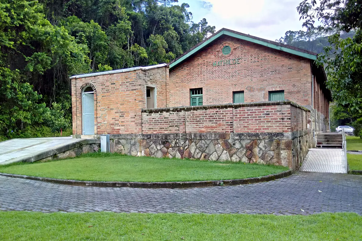 Prédio de tijolos da antiga estação ferroviária de Matilde, em Alfredo Chaves (ES), com gramado na frente e mata atlântica no morro ao fundo, em dia de sol