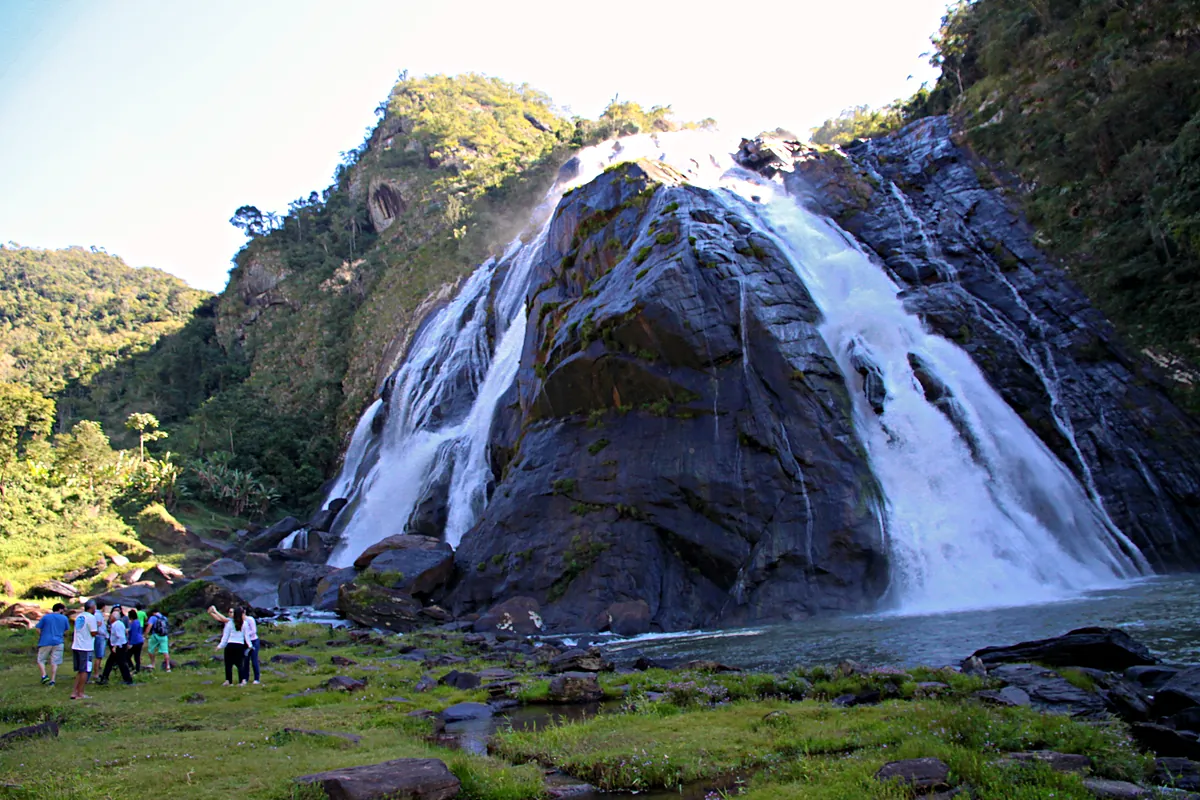 Cachoeira da Fumaça, em Alegre (ES), com a água caindo pela parede de pedra até o poço e um grupo de visitantes no gramado da margem, em dia de céu claro