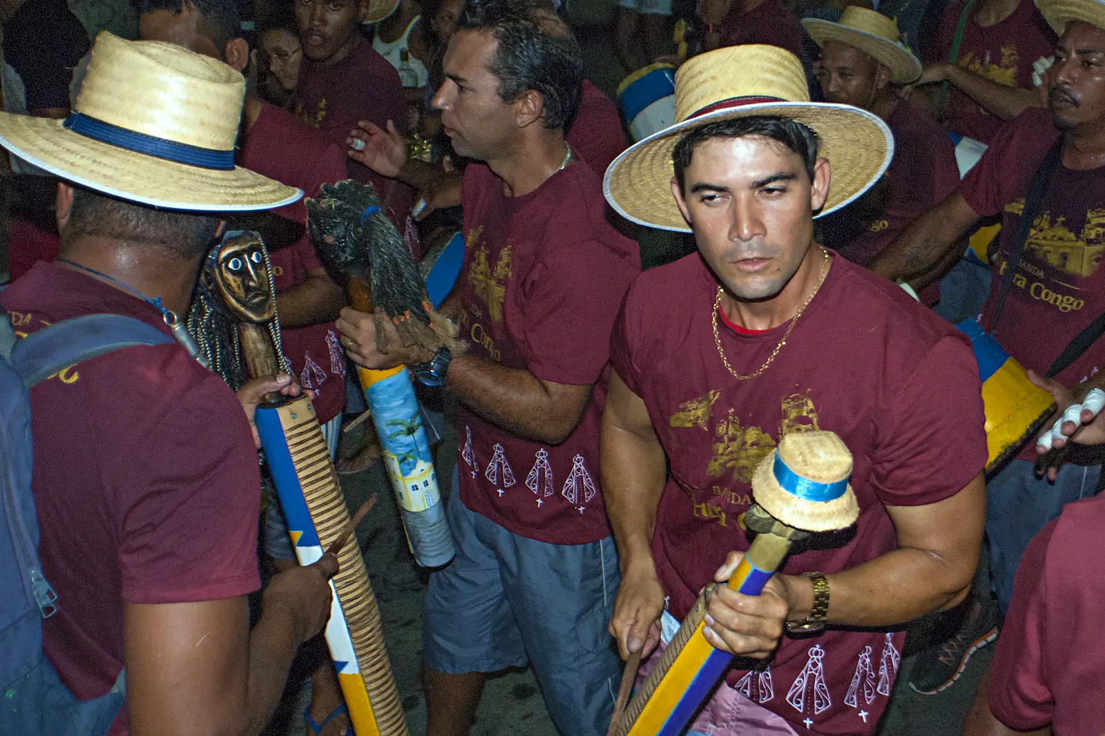 Tocadores de banda de congo na Serra (ES), de chapéu de palha e camiseta vinho, segurando casacas de cabeça esculpida durante festa à noite