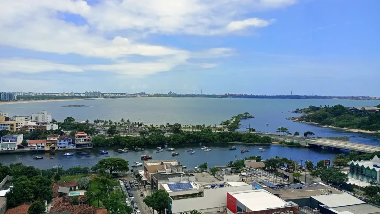 Vista do alto da Praia do Canto, em Vitória (ES), com o canal e barcos atracados, a ponte, a ilha arborizada e, ao fundo, a orla de Camburi sob céu azul com nuvens