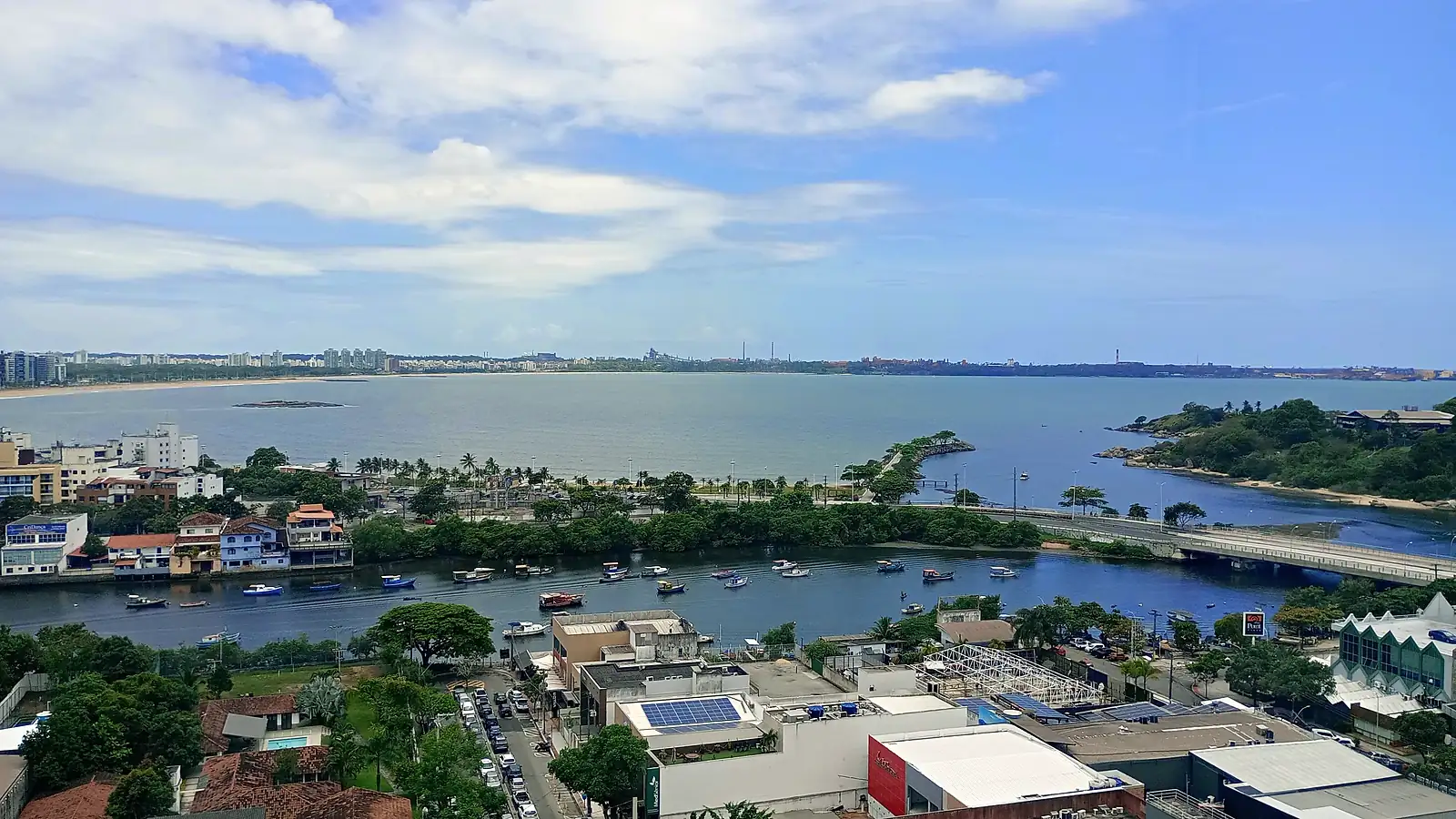 Vista do alto da Praia do Canto, em Vitória (ES), com o canal e barcos atracados, a ponte, a ilha arborizada e, ao fundo, a orla de Camburi sob céu azul com nuvens