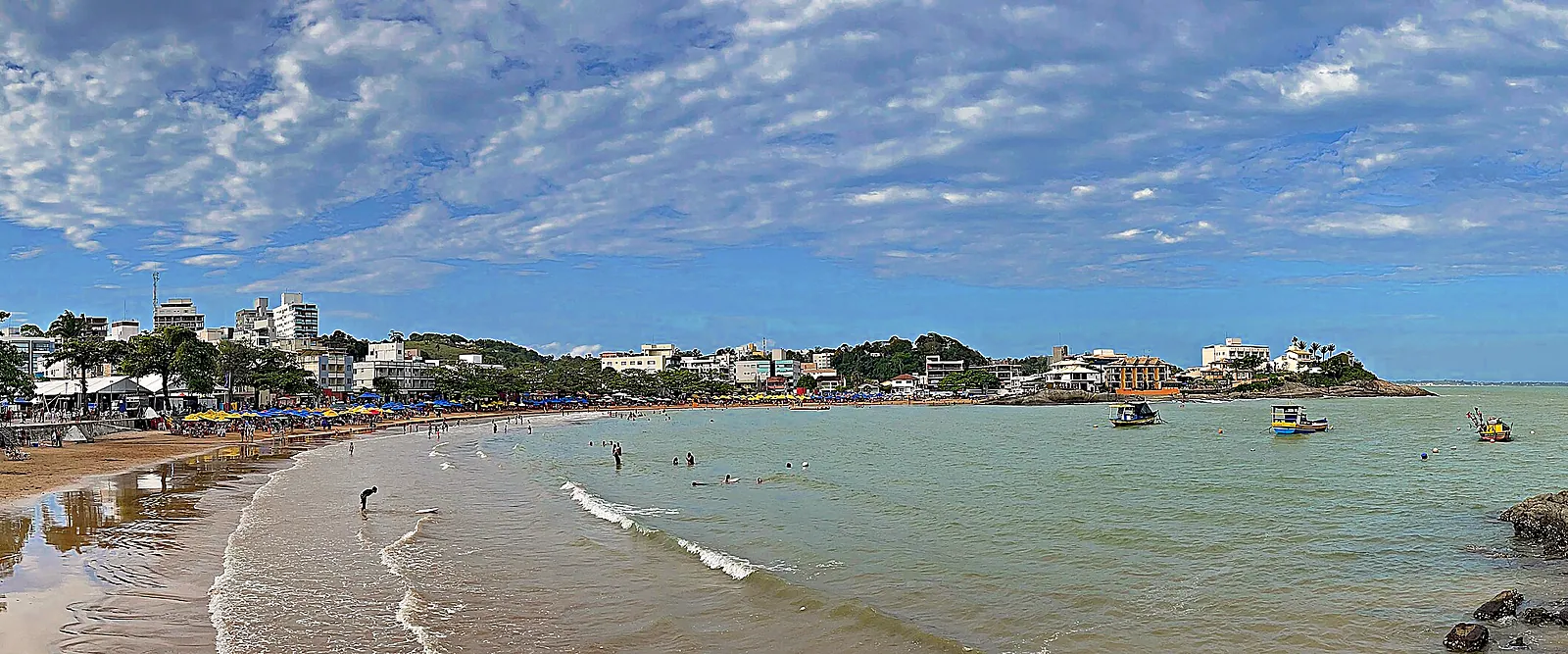 Orla de Iriri com árvores, prédios baixos, barcos ancorados e o mar calmo, vista do costão