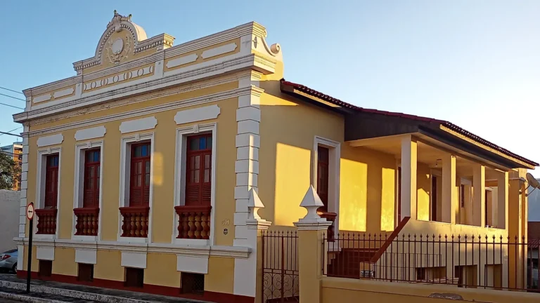 Casarão dos Calmons, sobrado amarelo com frontão branco ornamentado, janelas e balaústres vermelhos e varanda lateral com grade, no centro de Linhares (ES), com sol baixo na fachada e céu azul-claro
