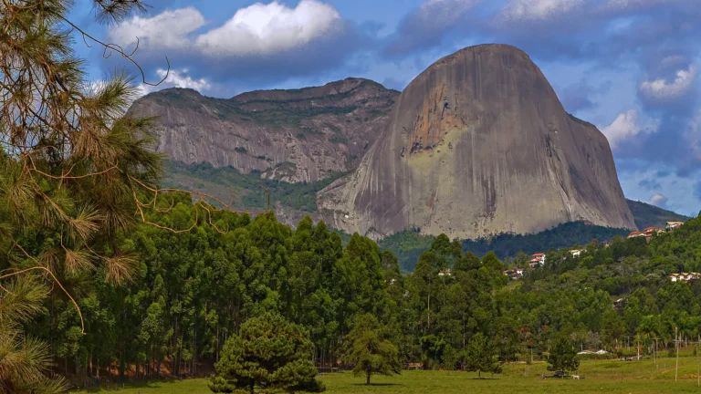 A Pedra Azul vista de um campo com pinheiros e eucaliptos, com casas na encosta à direita, em Domingos Martins (ES)