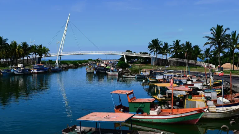 Barcos de pesca coloridos atracados no Rio Jacaraípe, perto da foz, na Serra (ES), com a passarela branca de pedestres sobre o rio, coqueiros nas margens e o mar ao fundo sob céu azul