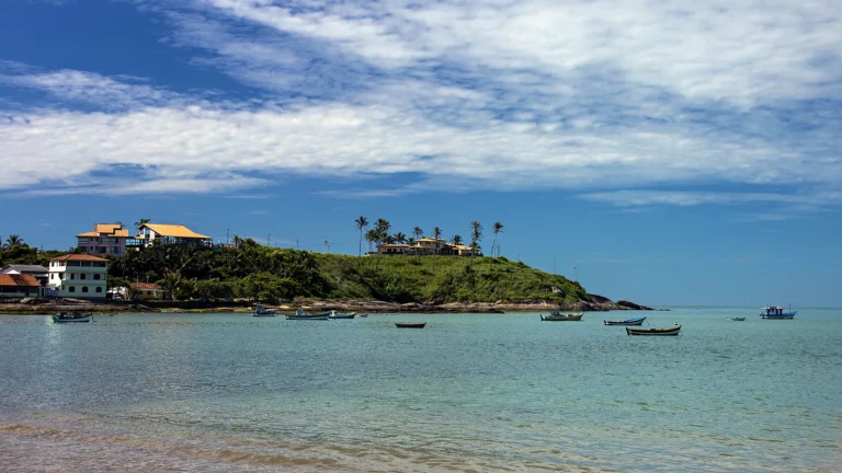 Barcos de pesca da Vila dos Pescadores ancorados na enseada de Meaípe, em Guarapari (ES), em 2018, diante da ponta verde com casas e coqueiros, sob céu azul com nuvens altas