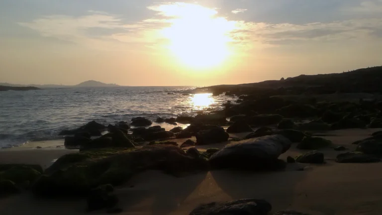 Praia do Ermitão ao nascer do sol, em Guarapari (ES), com pedras cobertas de musgo e areia em primeiro plano, o mar calmo refletindo o sol e o costão escuro à direita