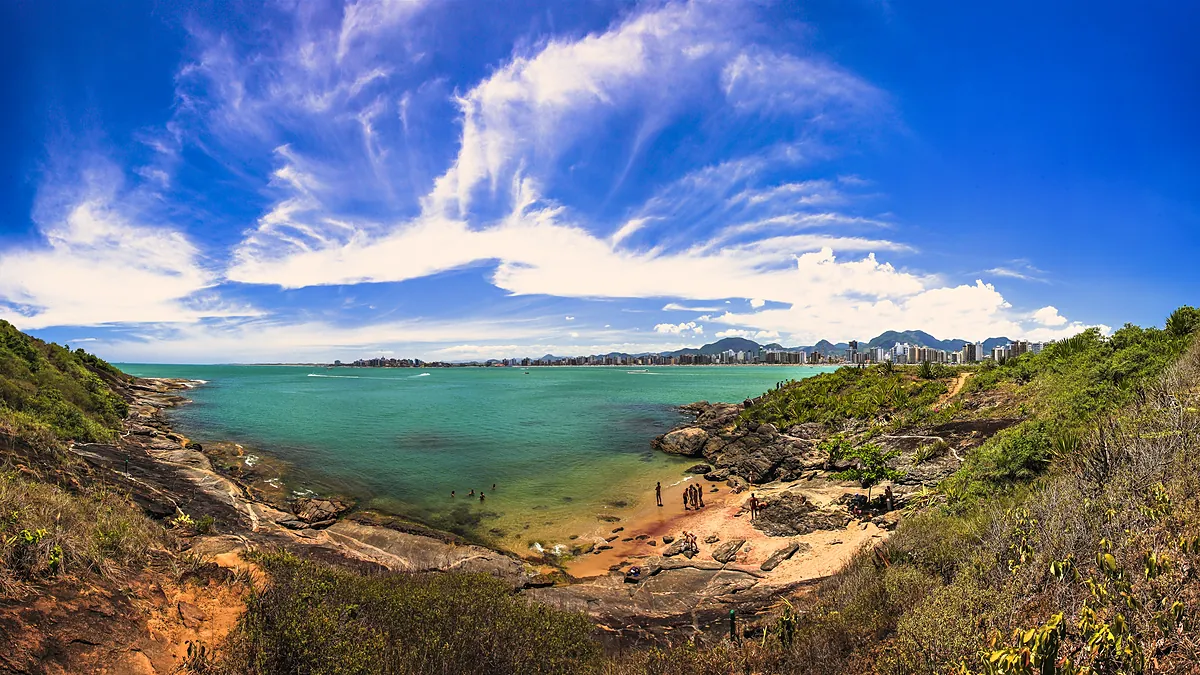 Enseada de mar verde-azulado vista do alto da trilha do Parque Natural Municipal Morro da Pescaria, em Guarapari (ES), com mata e pedras em volta e os prédios da Praia do Morro ao fundo