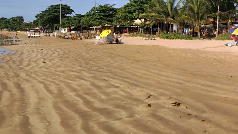 Orla da Praia Grande, em Fundão (ES), em 2010, na maré baixa, com a areia ondulada em primeiro plano, quiosques, amendoeiras e coqueiros ao fundo e céu azul