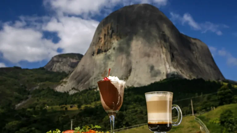 Taça de sobremesa de chocolate com chantili e cereja e copo de café em camadas ao ar livre, com a Pedra Azul e a mata ao fundo, em Domingos Martins (ES), em dia de céu azul