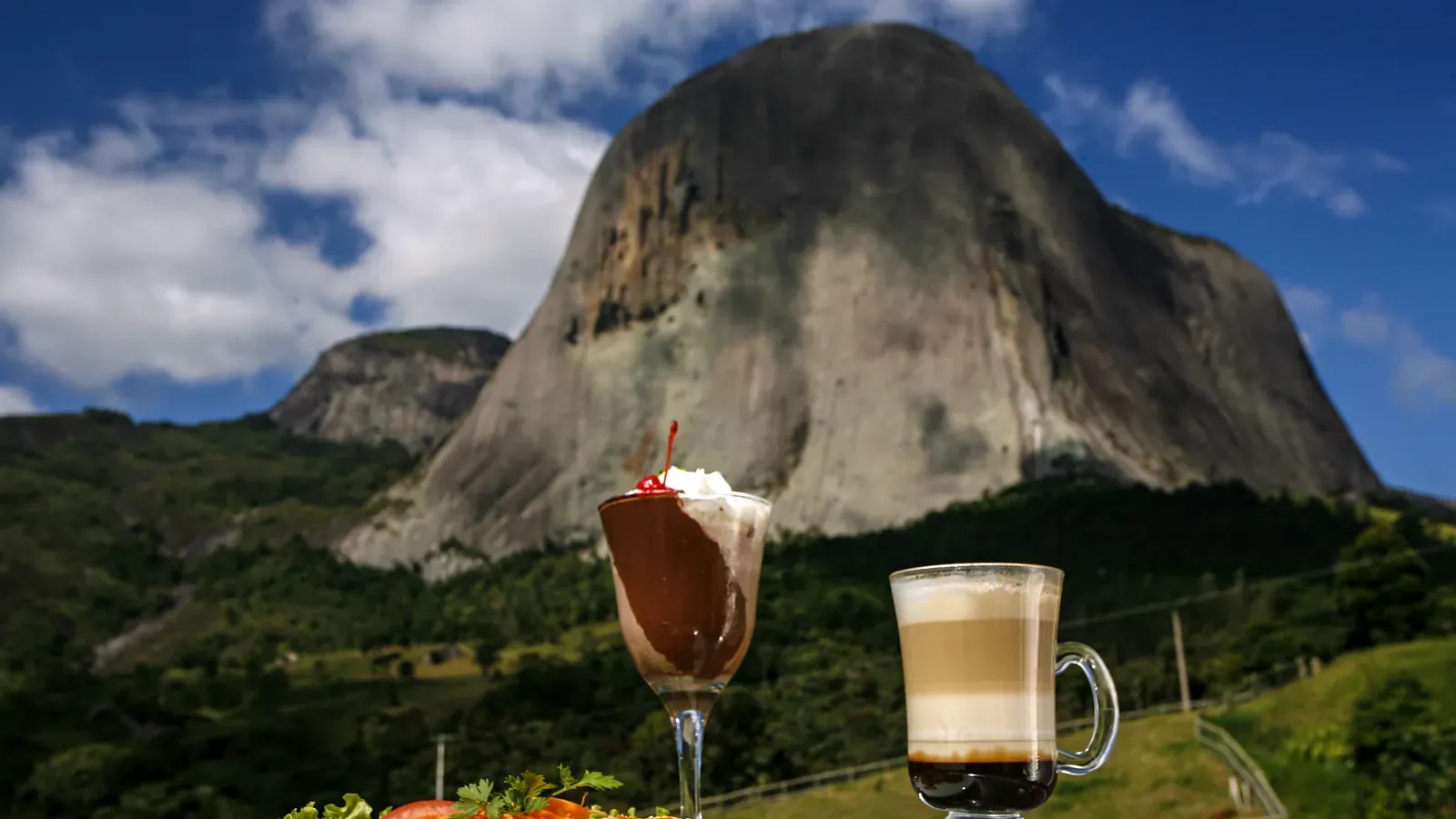 Taça de sobremesa de chocolate com chantili e cereja e copo de café em camadas ao ar livre, com a Pedra Azul e a mata ao fundo, em Domingos Martins (ES), em dia de céu azul