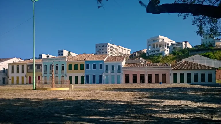 Casario colonial colorido do Sítio Histórico do Porto, em São Mateus (ES), com sobrados amarelos e azuis e uma casa térrea rosa diante do largo de pedra e terra e do pelourinho, sob céu azul limpo