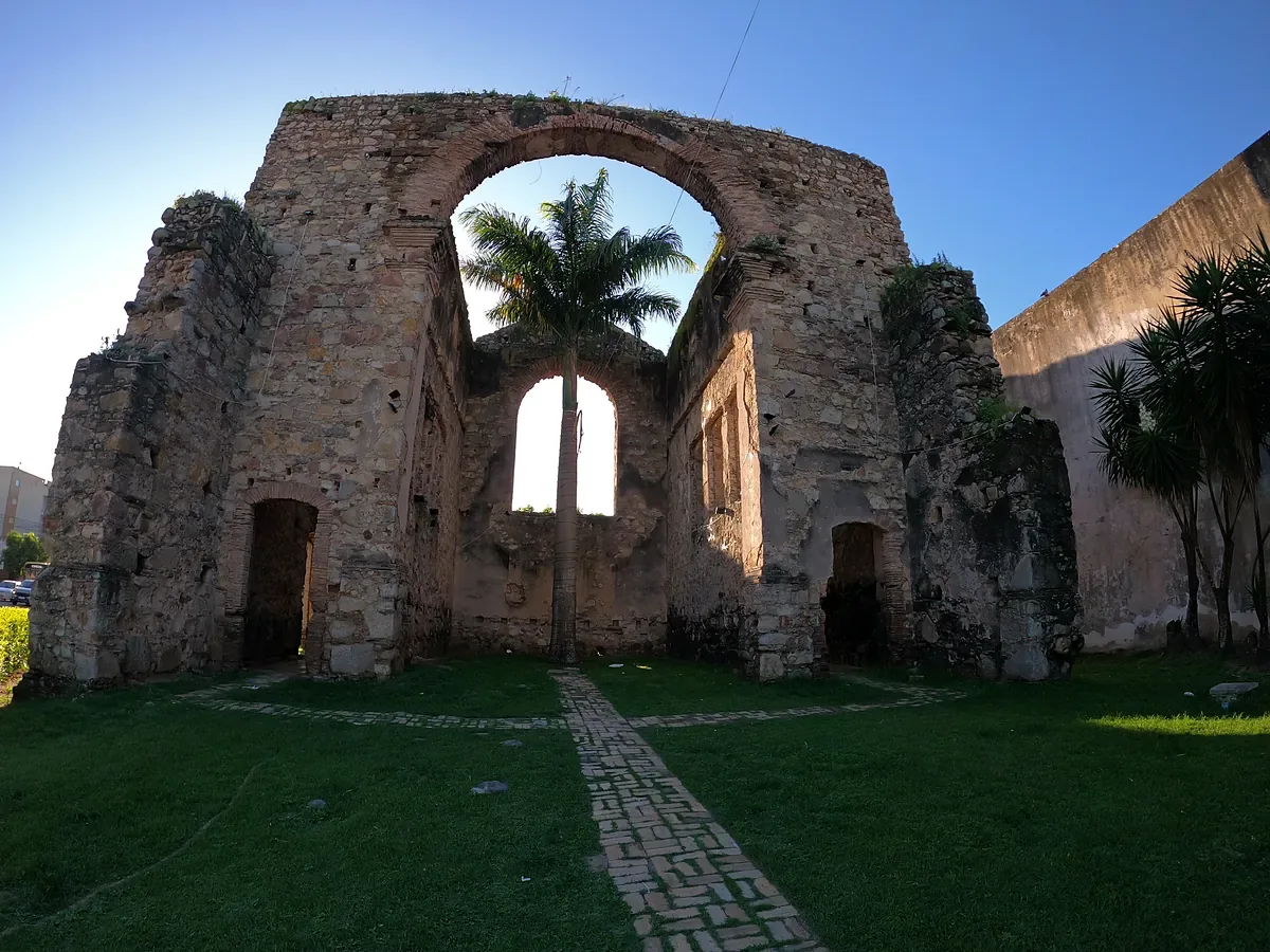 Ruínas de pedra da Igreja Velha no centro de São Mateus (ES), com o arco vazado da fachada, palmeiras atrás e gramado com caminho de pedra à frente, sob céu azul