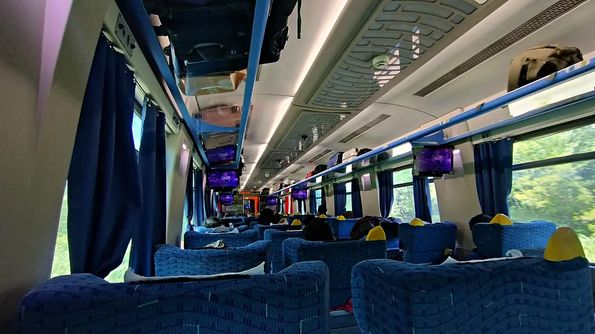 Interior de um carro da classe executiva do trem Vitória-Minas, com poltronas azuis de capa amarela no encosto de cabeça, cortinas azuis nas janelas, telas no teto e mata vista pelas janelas