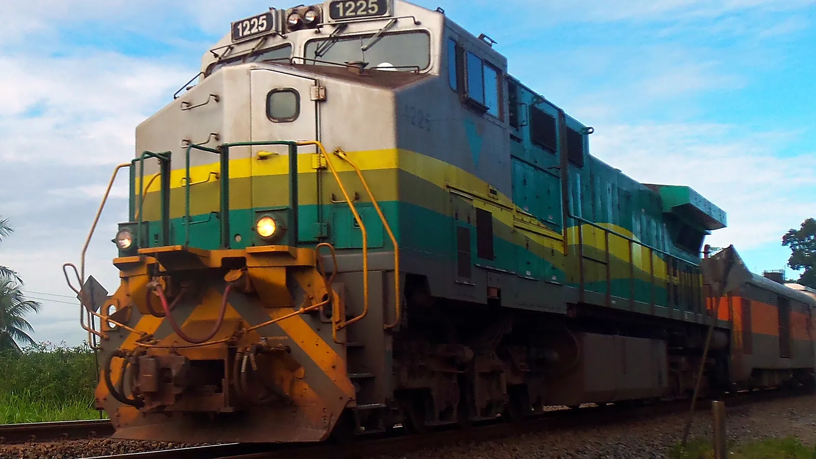 Locomotiva verde, amarela e cinza do trem de passageiros da Estrada de Ferro Vitória a Minas vista de baixo, com um carro de passageiros atrás e céu azul com nuvens