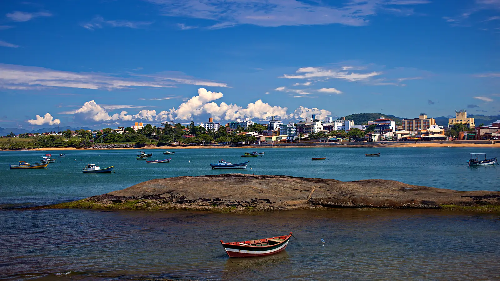 Praia de Meaípe em Guarapari (ES) com barcos de pesca ancorados na água calma, uma laje de pedra no meio e a fileira de prédios baixos da orla ao fundo sob céu azul