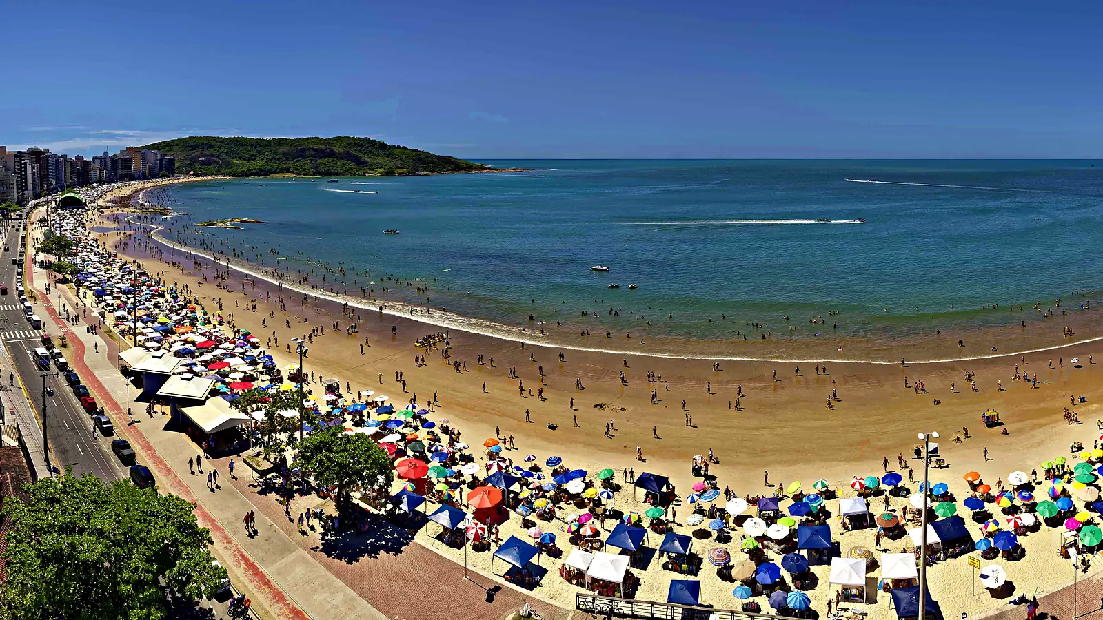 Praia do Morro em Guarapari (ES) vista do alto num dia de sol, com a faixa de areia cheia de guarda-sóis, a avenida da orla à esquerda e o morro coberto de mata ao fundo